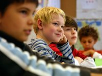 Three young boys are sitting in class, looking towards the front of the room, smiling. 