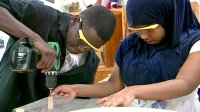 Wearing safety goggles, a boy working a drill while a girl steadies the project using two hands