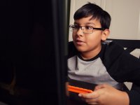 A young boy is sitting, looking at a screen, with an orange keyboard in his hands.