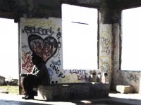 A man is sitting in an abandoned building with graffiti on the cement walls and benches. 