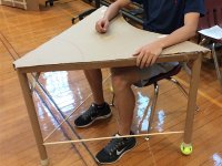 Chest down, a teen is sitting at a hand-made desk. It's made of a cardboard triangular desktop, three cardboard tubes as desk legs -- with tennis balls at the bottom of two of them -- and two solid, cylindrical wooden rods connecting the desk legs.