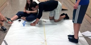 A teenage boy is kneeling on the floor in a school hallway beside a large piece of paper. He's drawing straight lines on it against a metal ruler. A teenage girl is sitting beside him, cutting something with scissors. Two other teenagers are watching.