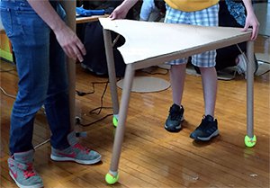 Two students are holding up a cardboard-made desk. The desk looks wobbly with two legs off-center. 