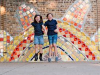 Two girls jumping up in front of a large tiled wall of a sun