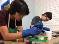 Two teenage boys in protective goggles, green aprons, and blue gloves are hovering over a desk holding a screwdriver, using it with some plastic-metal device.