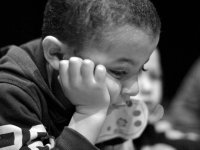 A black and white closeup of a young boy, maybe 5-years-old, in a hoodie. He's leaning forward with his head resting on his hand, and he has a solemn look on his face. 