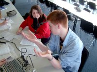 Teenage students are sitting at a long table in a classroom. One of them is talking into a microphone. They have a soundboard next to them with papers scattered across the table. 