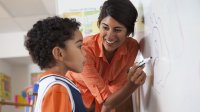 A teacher works at the white board with a young boy. 