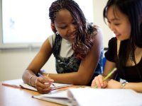 Two older girls side by side at desk looking at binders speaking with each other