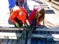 Two young students are outside, bending over planks of wood, hammering them together.