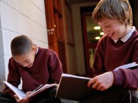 Two boys sitting in the hallway writing in their notebooks, talking, and smiling