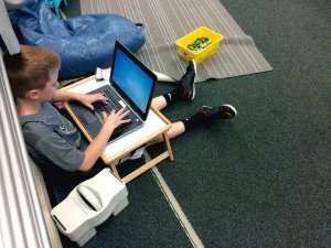 A young boy typing on a laptop on a lap desk while sitting on a classroom floor