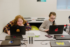 Two girls type on laptops in their classroom.
