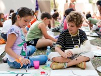 A group of young students are painting on paper, sitting outside on large pieces of cardboard laid out on the grass.