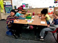 Four young students are sitting together at two, two-person desks put together as one table. They're near the front of the classroom near a whiteboard. The two girls are looking down, writing, and the two boys are looking up, smiling.