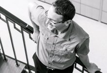A male teacher in glasses is leaning against a stairway rail, smiling. 