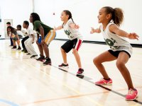 Seven young students are squatting side-by-side on a basketball court with their arms extended out at their sides.