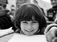 A closeup of a young kid's face, smiling, with their chin perched on their desk next to an open textbook.