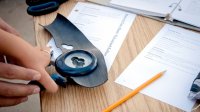 An adult's hands are guiding a student's hand to use an upscale hole punch. They're holding it on top of a desk. There's an opened binder, papers, scissors, and a pencil strewn across the desk.