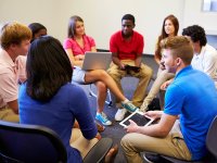 In the middle of a classroom, a group of high school students are sitting in chairs in a circle, talking to each other.
