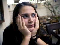 A closeup of a teenage girl sitting slouched, resting her head in her cupped hands. She's looking up and to the side with tears in her eyes. She's sitting in a naturally lit room with couches, comfy chairs, coffee tables, and a small, potted tree.