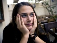 A closeup of a teenage girl sitting slouched, resting her head in her cupped hands. She's looking up and to the side with tears in her eyes. She's sitting in a naturally lit room with couches, comfy chairs, coffee tables, and a small, potted tree. 