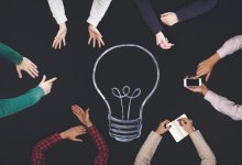 Top view of six pairs of hands around a chalk drawing of a lightbulb.