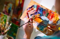 A close-up of a person holding a wooden palette with a variety of acrylic paints on it