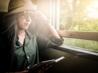 A woman in sunglasses and a hat sitting on a train on a sunny day with the window cracked open, reading a book