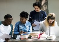 A group of four teenagers reading and discussing literature together on an outdoor table at school