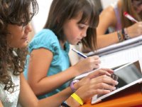 A girl writing in a binder and sitting next to her, a girl on a tablet