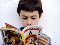 A closeup of a young boy in a blue striped shirt against a white wall reading a book