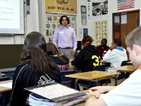 Six high school students are sitting at individual desks, looking towards the teacher at the front of the class.