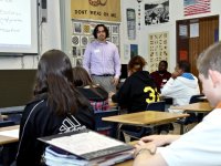 Six high school students are sitting at individual desks, looking towards the teacher at the front of the class. 