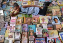A young black girl is laying down on top of about 300 books stacked together on top of each other, looking up at the camera, smiling.