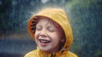 A young boy in a yellow rain coat is smiling with his eyes closed, and rain is pouring down on him.