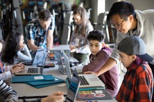 Group of middle school students working on laptops with teacher supervision.