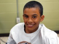 A closeup from the chest up of a young boy smiling directly at the camera. He's wearing a white t-shirt, and behind him is a green wall. 