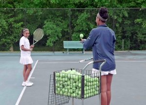 A teenage girl and younger girl are smiling, standing outside on a tennis court. The older girl is throwing a tennis ball to the younger girl.