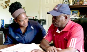 A teenage girl and a man are sitting at a table in a house, both looking at an opened textbook.