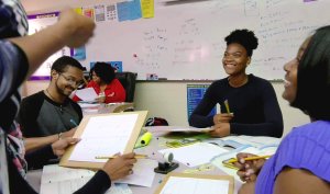 Four students are sitting at a classroom table smiling. One of them -- a teen girl -- is looking up, smiling at someone who is standing outside of the photo.