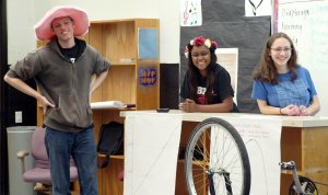 A tall teenage boy in a pink hat is standing at the front of the class with his hands on his hips. He's standing beside two teenage girls who are behind a desk turned into a podium. The three of them are smiling out to an audience.