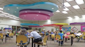 Young students are working at tables in a large multipurpose room with large colorful-shaped decorations hanging from the ceiling. 