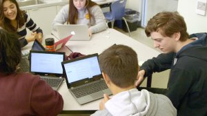 Five high school students are sitting at two tables pushed together. They are working together and talking with their laptops opened.