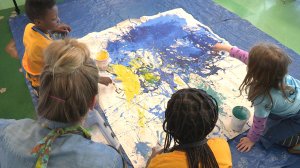 Three young students and a teacher are kneeling on a blue tarp in class pouring paint onto a large canvas.