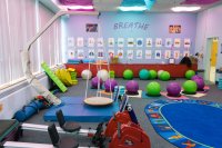 A colorful classroom with light purple walls, blue and purple cloths covering the ceiling lights, a swing set, bouncy balls to sit on, and rowing exercise equipment.