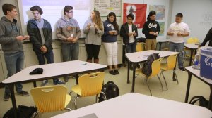 Eight high school students are standing in a row at the front of the class, each ripping a small piece of paper.