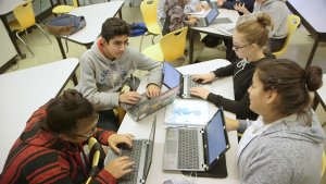 Four high school students are talking to each other, sitting at a trapezoidal table with laptops in front of each of them.