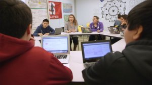 High school students are sitting in a circle made of trapezoidal desks at the center of the room, while another student works by herself, facing the wall.