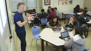 A teacher is standing by a projector talking to three students who are sitting at two small tables pushed together. A group of six students are working together at the side of the room, and three students are working by themselves at the back of the room.